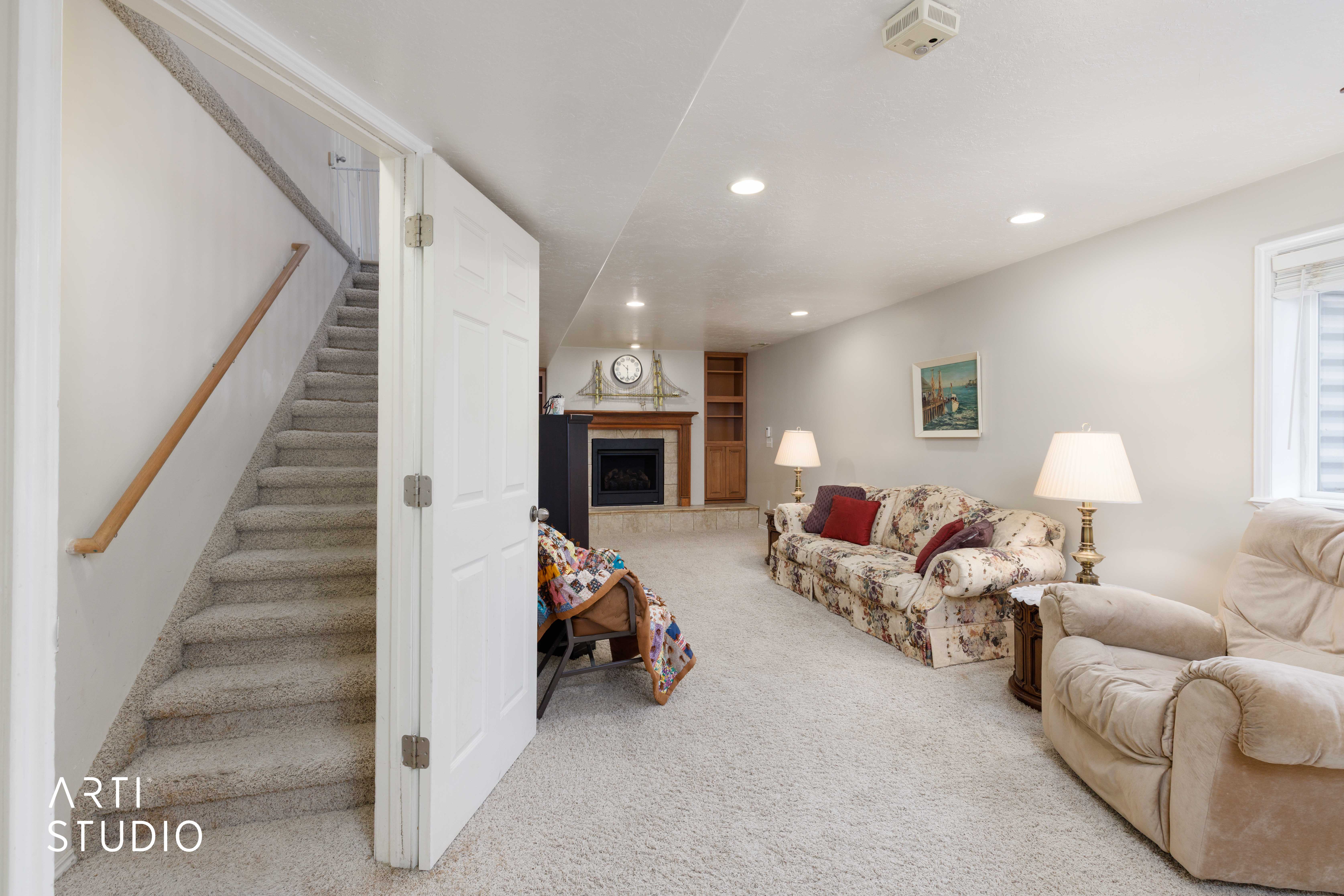 Carpeted lower-level living area featuring a fireplace with a wood mantel, recessed lighting, built-in shelving, a window with horizontal blinds, and a staircase with a wood handrail