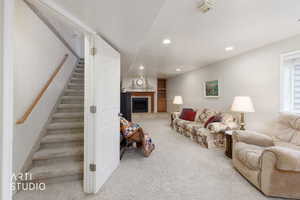 Carpeted lower-level living area featuring a fireplace with a wood mantel, recessed lighting, built-in shelving, a window with horizontal blinds, and a staircase with a wood handrail