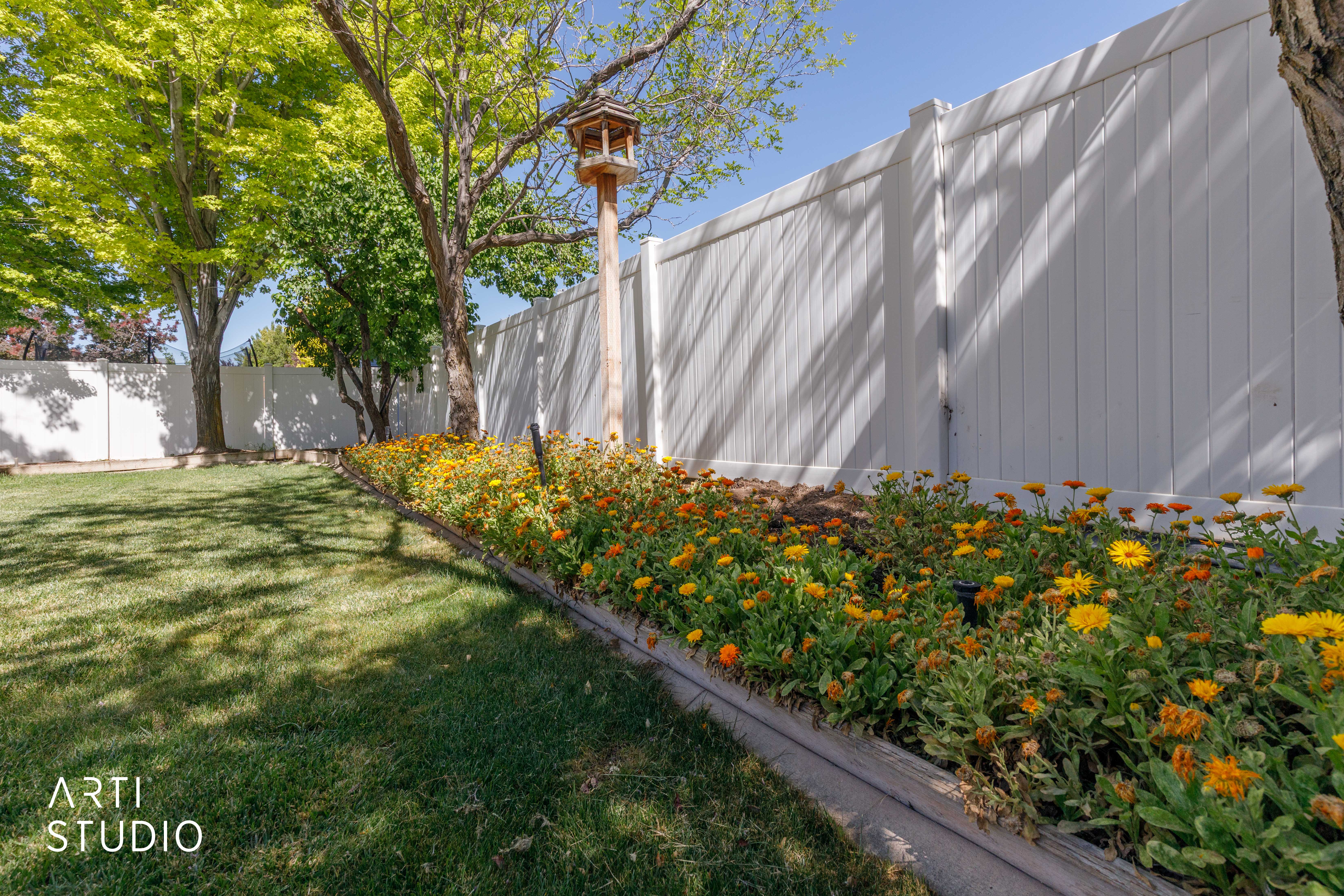 Expansive lawn area featuring a garden bed with blooming yellow and orange flowers, mature trees, and a white privacy fence