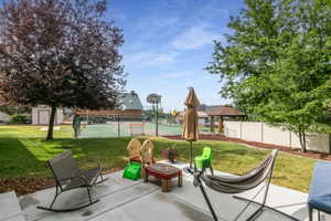 Expansive backyard featuring a concrete patio, a manicured lawn, mature trees, a sports court with fencing, and a detached storage shed.