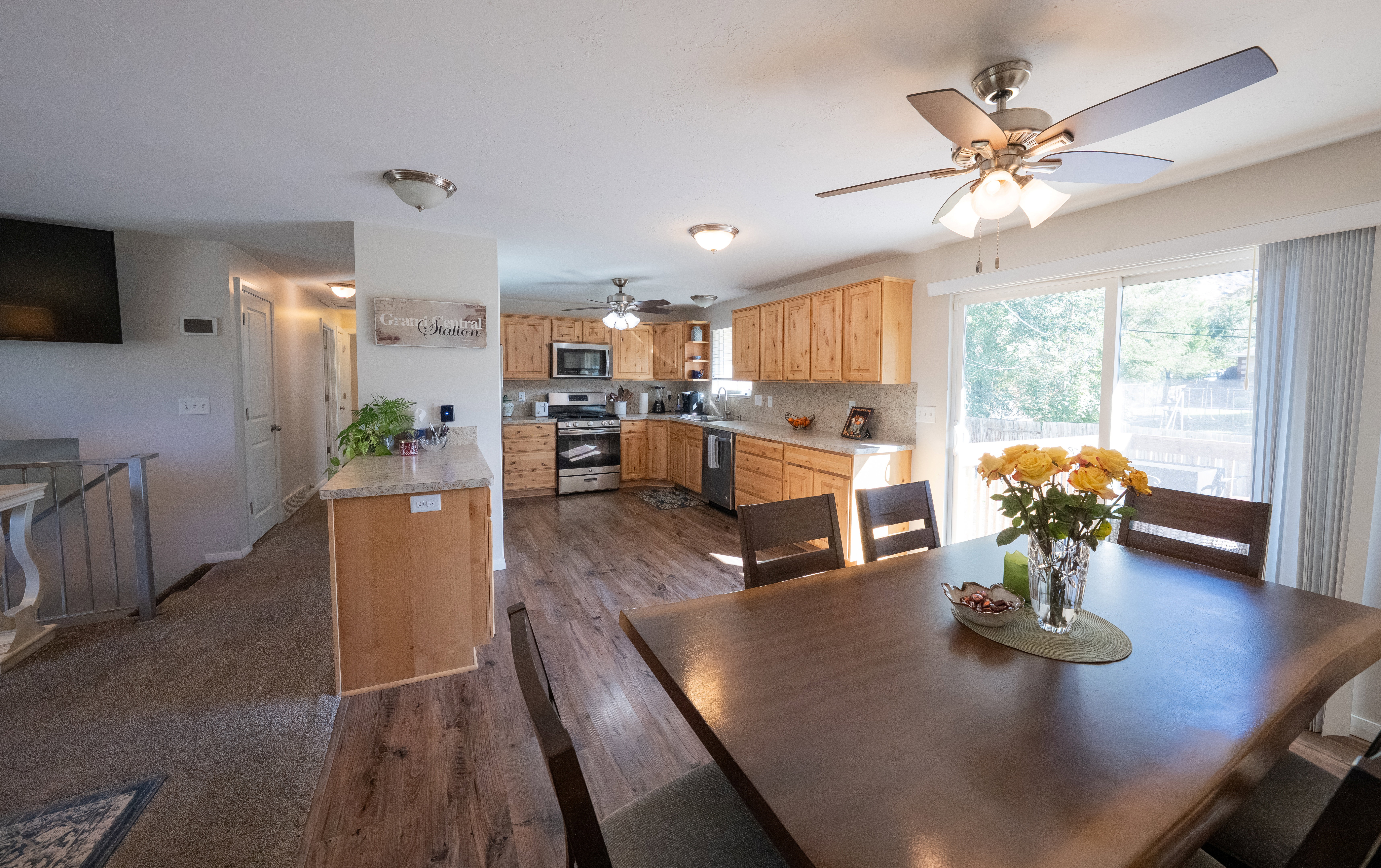Open-concept living space featuring wood-finish flooring, a kitchen with light wood cabinetry and light-toned countertops, stainless steel appliances, a tile backsplash, and a sliding glass door