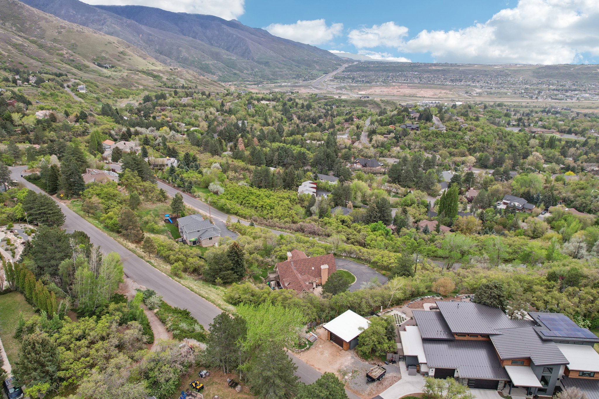 Expansive aerial showcasing a mountainous backdrop, verdant hillside properties, a winding paved road, a contemporary multi-level residence with a dark roof and solar panels, and abundant mature trees