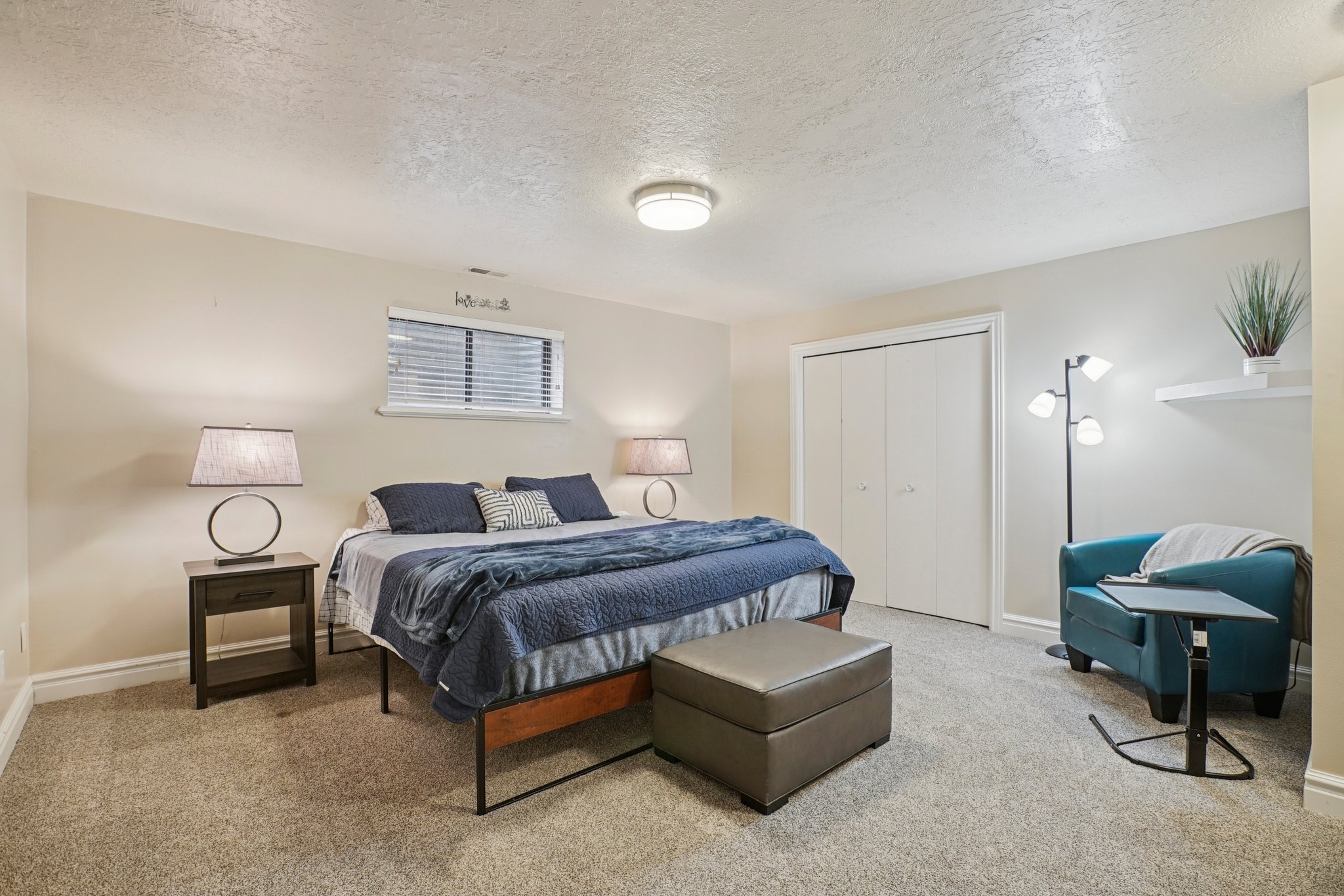 Carpeted sleeping area featuring a window with horizontal blinds, built-in bi-fold closet doors, and a wall-mounted shelf