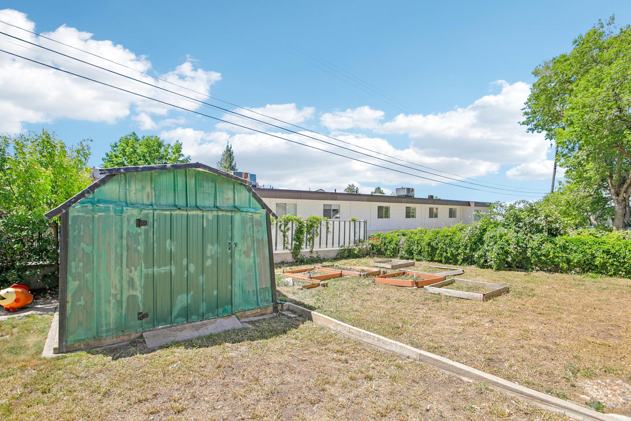Expansive backyard featuring a green utility shed with a gambrel roof, multiple raised garden beds, mature perimeter shrubbery, and a concrete border