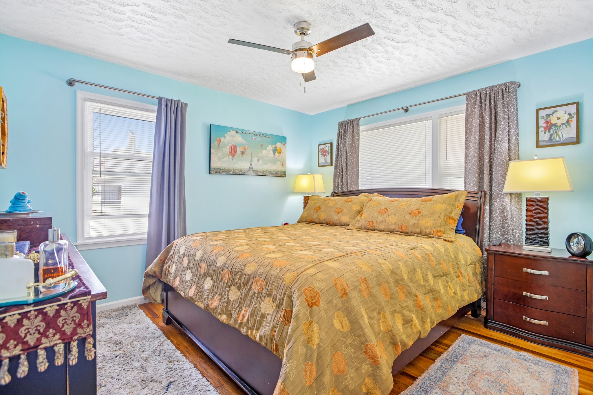 Bedroom featuring two windows with blinds and drapes, a ceiling fan with integrated lighting, and wood-finish flooring partially covered by an area rug