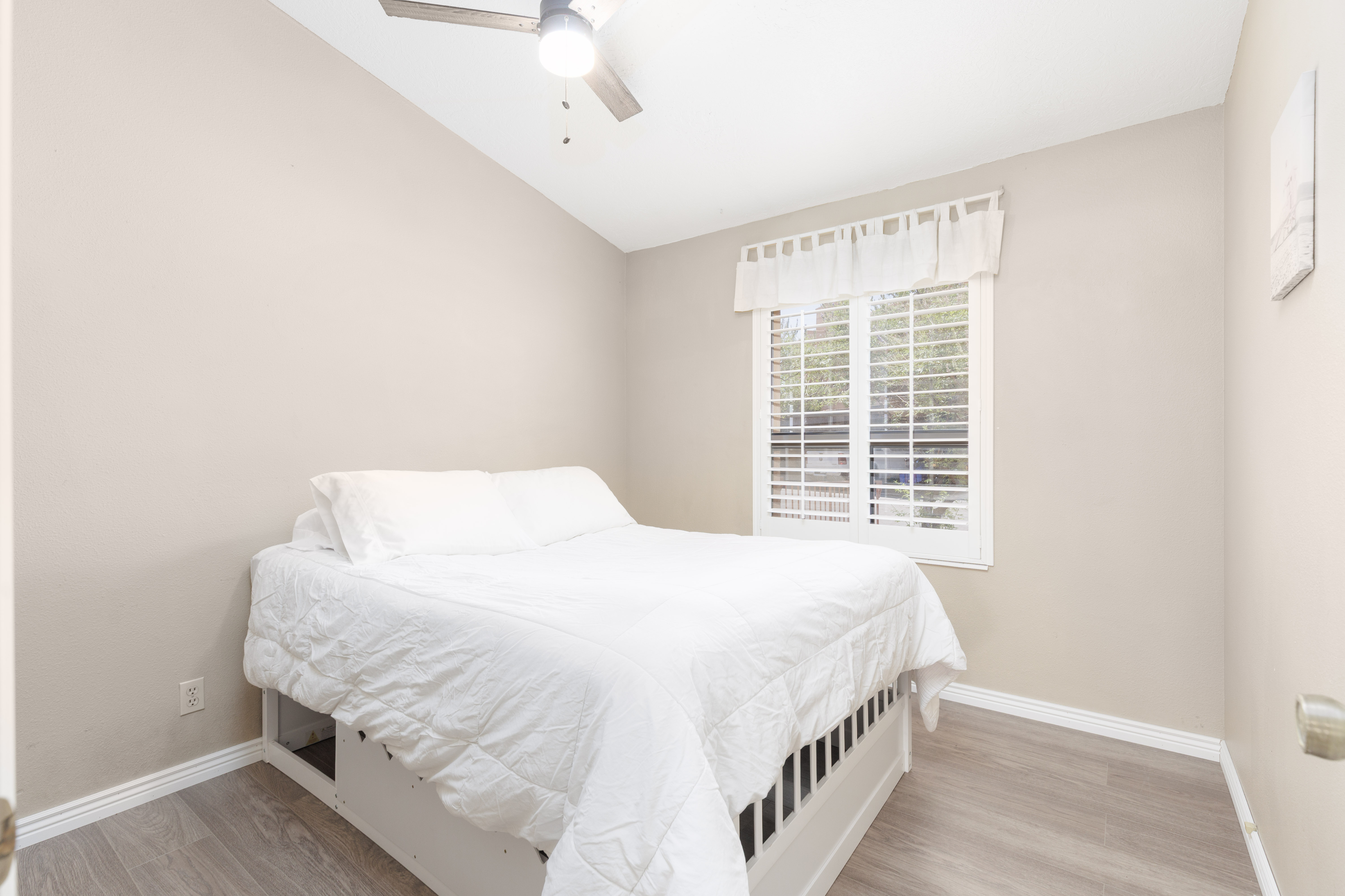 Vaulted ceiling with a ceiling fan light fixture, window with plantation shutters, wood-finish flooring, and light neutral wall paint