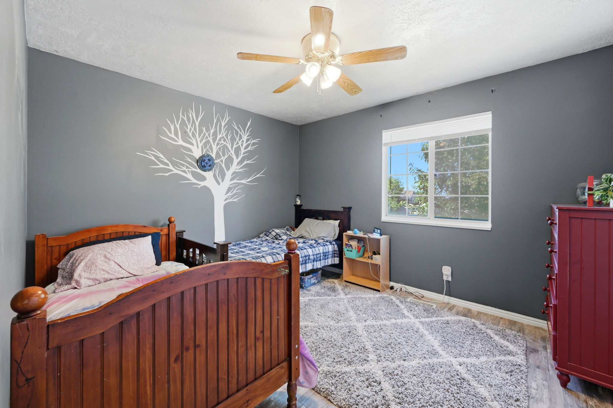 Spacious room featuring a ceiling fan with integrated lighting, a large window providing natural illumination, wood-finish flooring, and a decorative wall decal