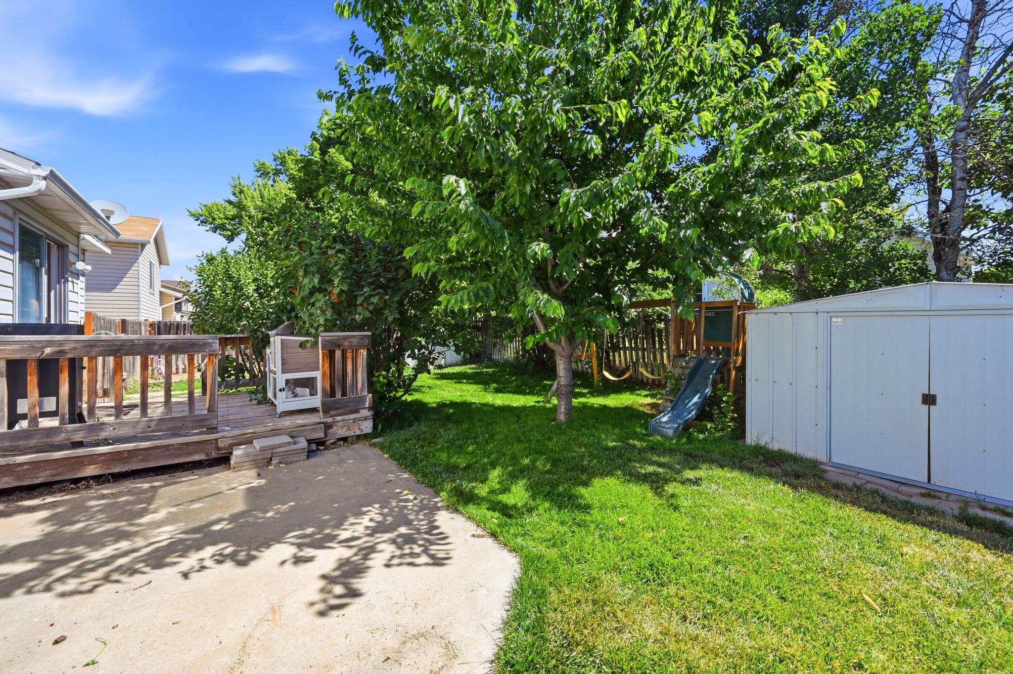 Expansive backyard featuring a concrete patio, a raised wooden deck, a mature deciduous tree, a metal storage shed, and a wooden playset with a slide and swings