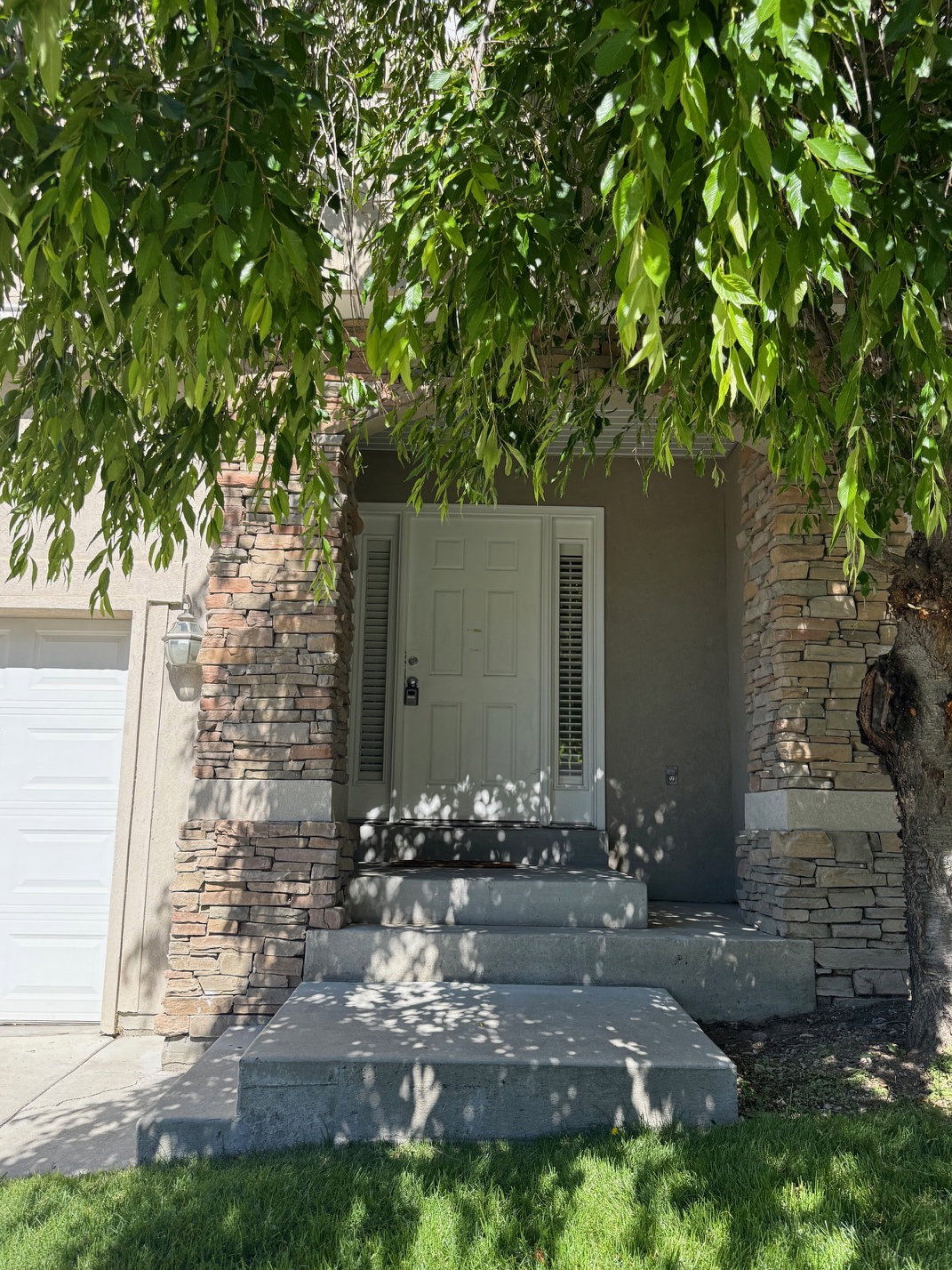 Front entry featuring a paneled door with sidelights, stacked stone accents on architectural columns, a stepped concrete walkway, and a garage with a white sectional door