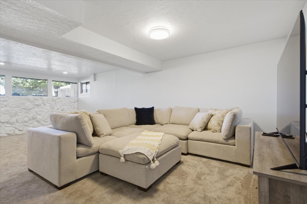 Spacious lower-level room featuring light-toned carpeting, white painted walls, a textured white stone wall, and multiple horizontal windows