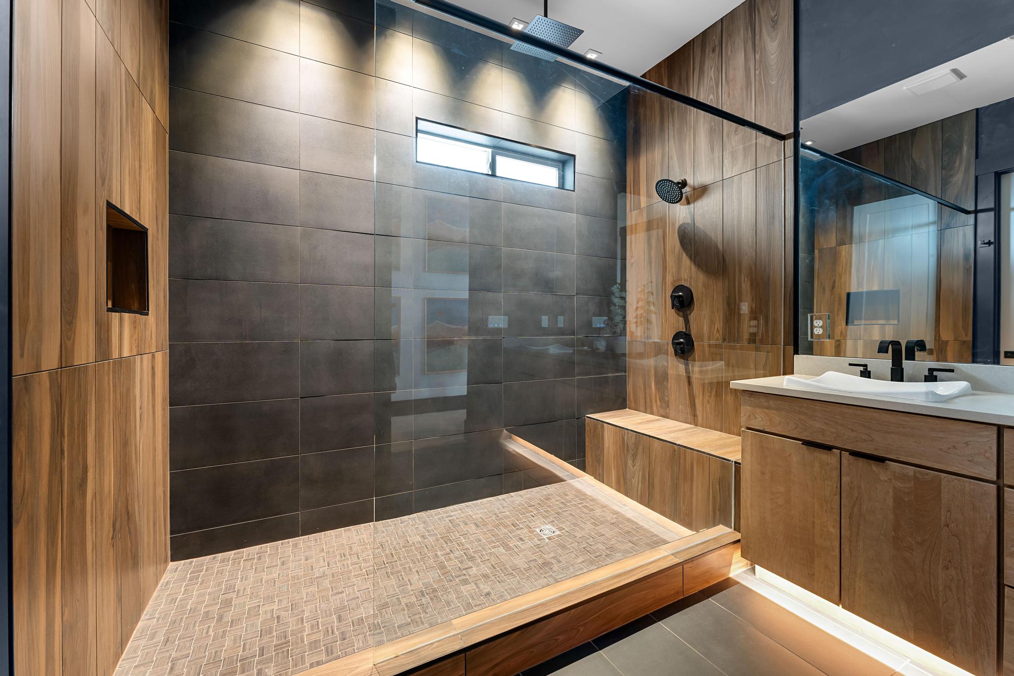 Spacious shower enclosure with wood-finish paneling, dark large-format tile, glass partition, and integrated bench seating