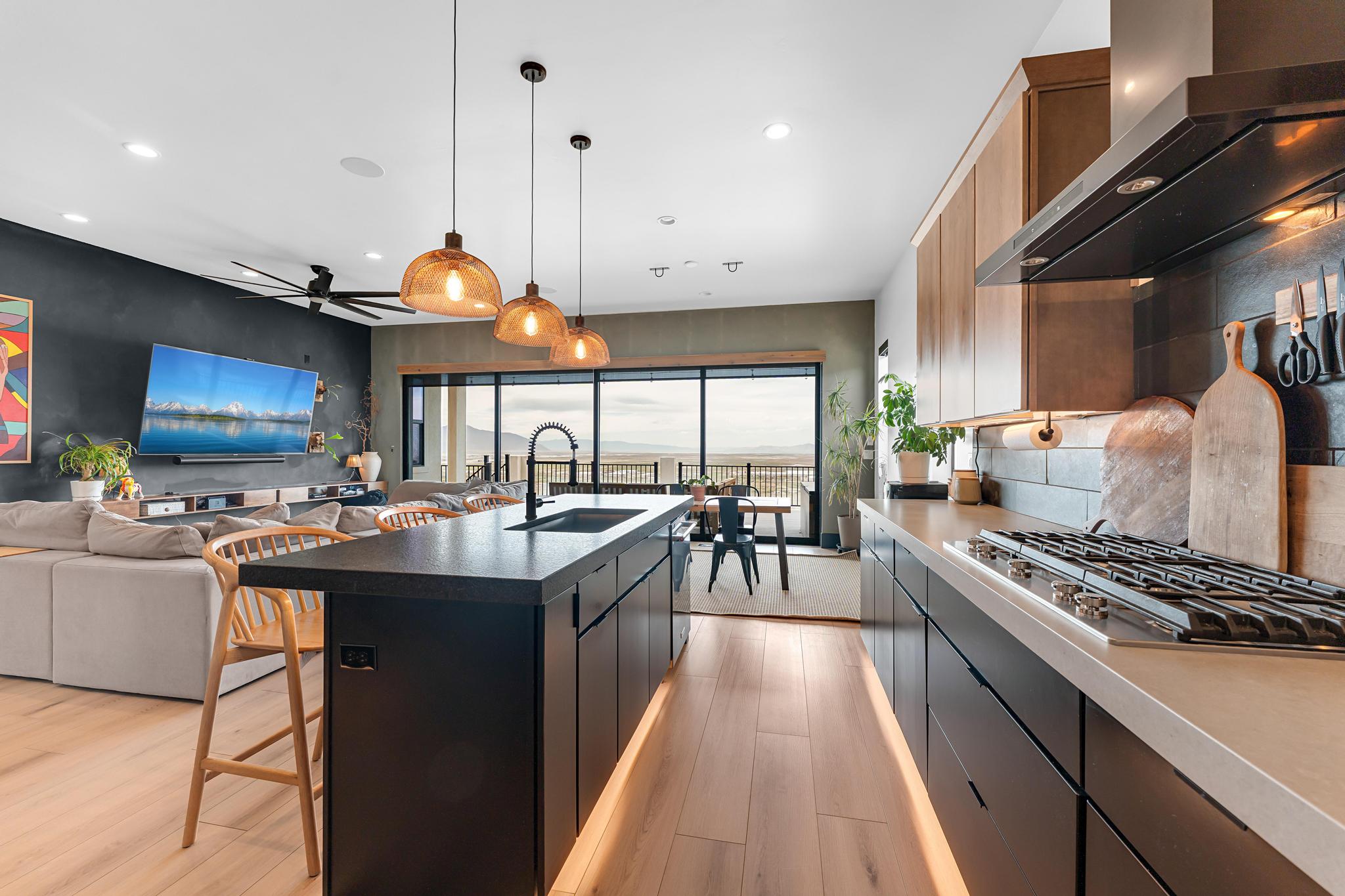 Open-concept kitchen featuring a dark island with integrated sink, pendant lighting, hardwood-finish flooring, and a gas cooktop with modern hood