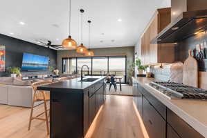 Open-concept kitchen featuring a dark island with integrated sink, pendant lighting, hardwood-finish flooring, and a gas cooktop with modern hood