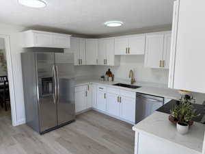 Modern kitchen featuring white shaker cabinetry, light-toned wood-finish flooring, white countertops, a stainless steel refrigerator, and a stainless steel sink with a gold-tone faucet
