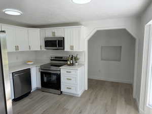 Kitchen featuring white shaker cabinetry with gold-finish hardware, stainless steel appliance suite, light-toned wood-finish flooring, and white countertops