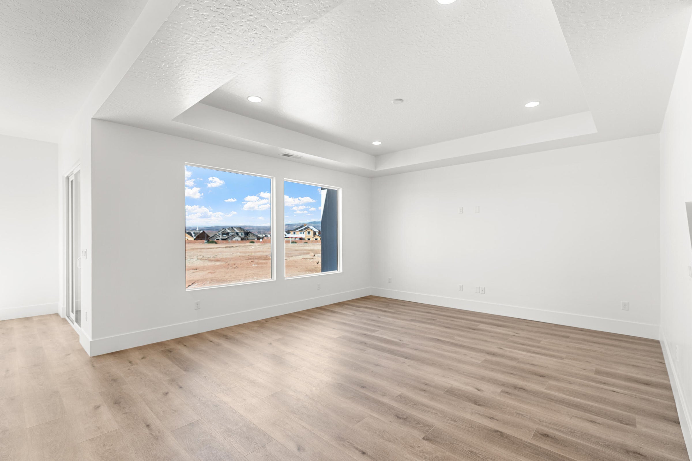 Spacious room featuring wood-finish flooring, a tray ceiling with recessed lighting, and two large windows offering distant scenery