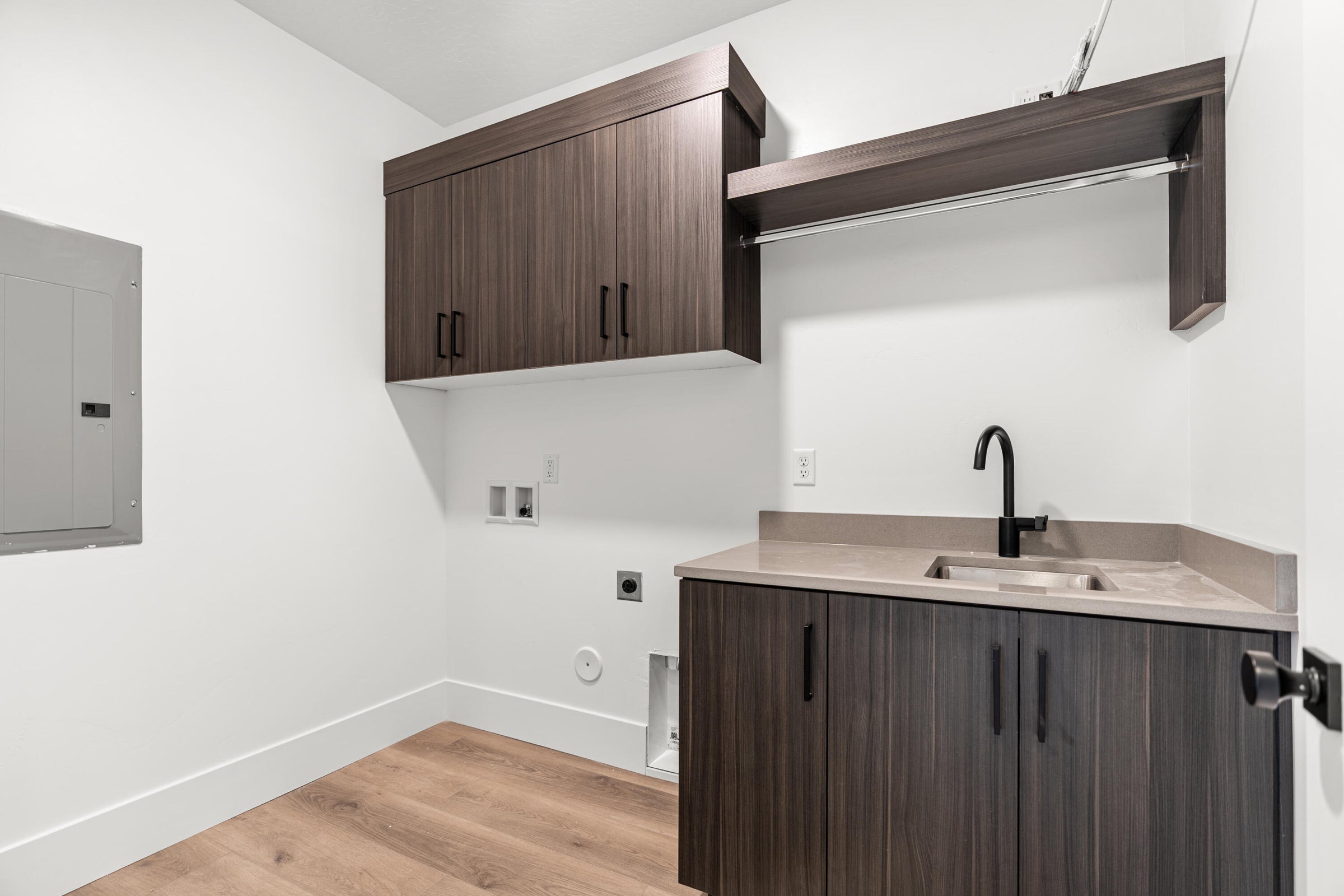 Laundry area featuring wood-finish flooring, a light gray countertop with an integrated sink, matte black faucet, and dark wood-finish cabinetry