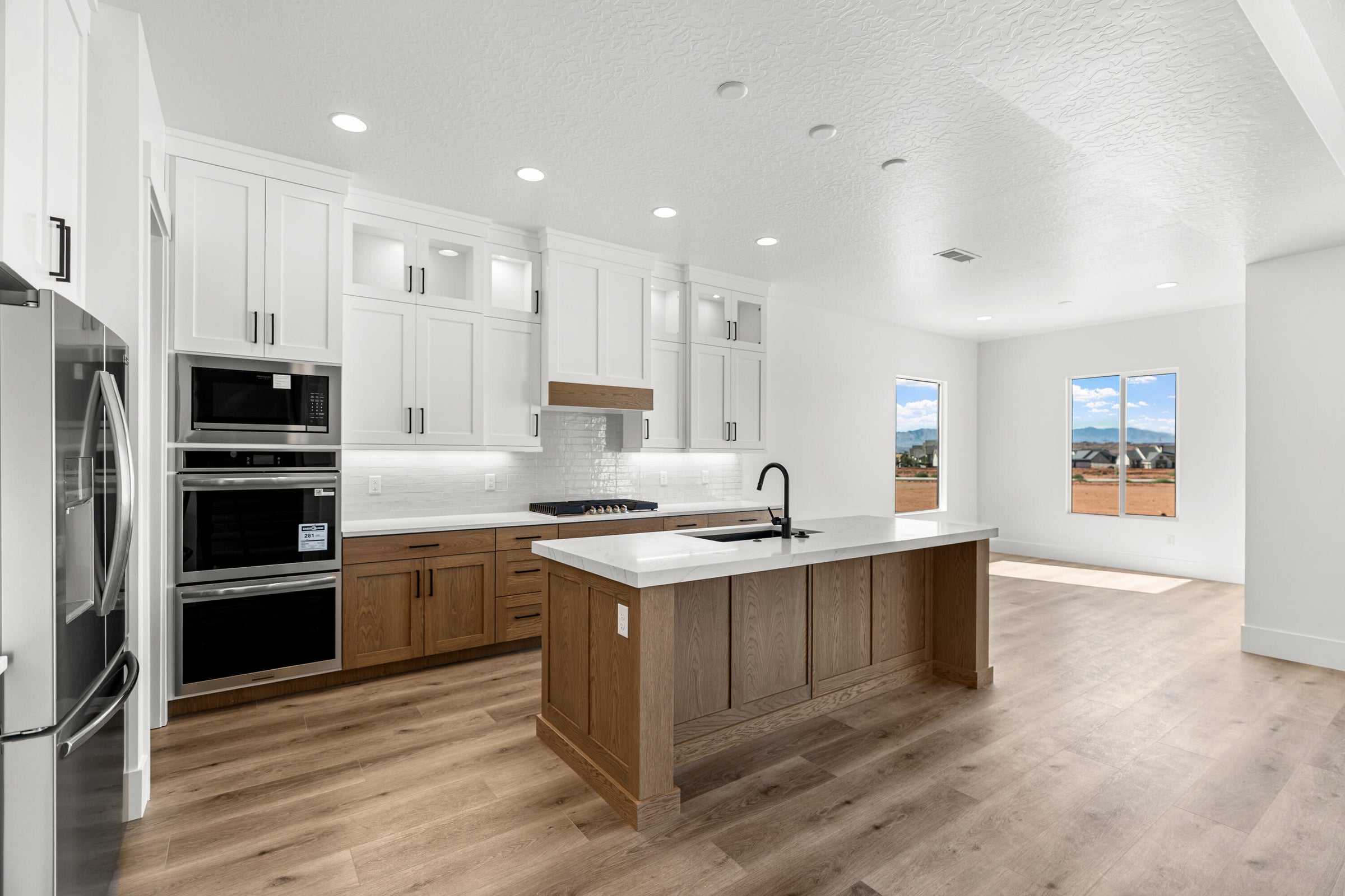 Open-concept kitchen featuring an oversized island with a light-toned countertop, white upper cabinetry, natural wood-finish lower cabinetry, and stainless steel appliances including a built-in microwave and double oven