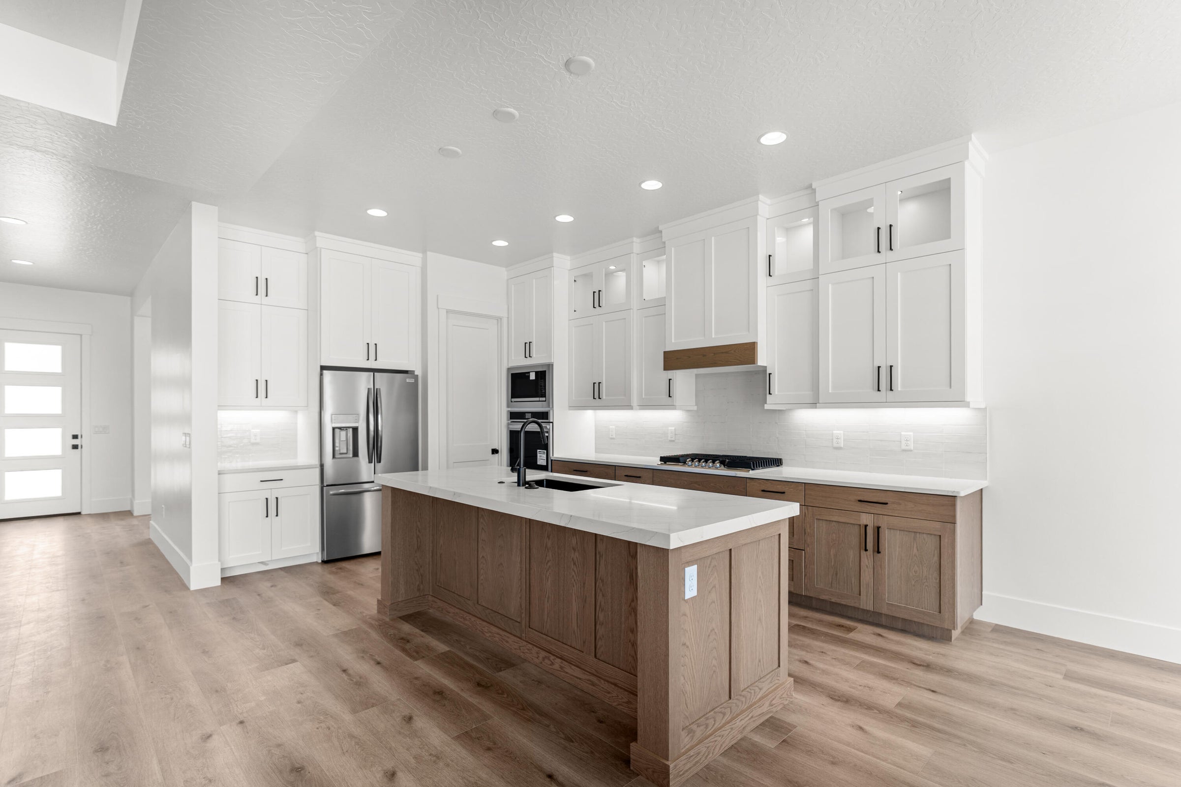 Contemporary kitchen featuring a large central island with a solid surface countertop, wood-finish flooring, white upper cabinetry with under-cabinet lighting, natural wood tone lower cabinetry, and stainless steel appliances