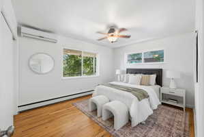 Bright room featuring two windows, wood-finish flooring, a ceiling fan with integrated lighting, and a wall-mounted climate control unit