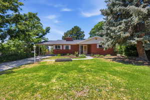 Red brick exterior residence featuring a front lawn with established trees, a curved concrete pathway, a covered carport, and a prominent chimney