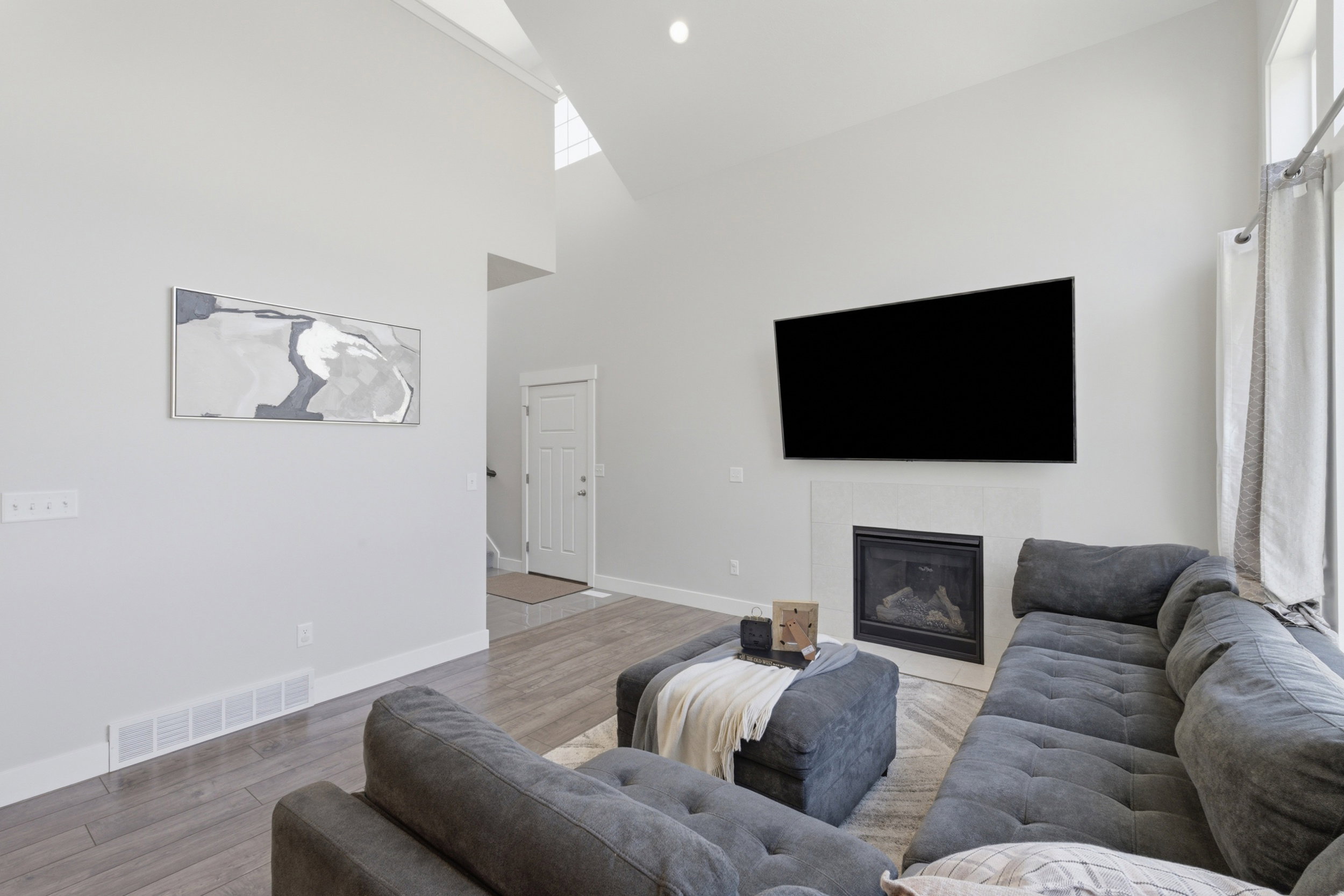 Living area featuring vaulted ceilings, a fireplace with a light-colored surround, wood-finish flooring, and a large window with drapes