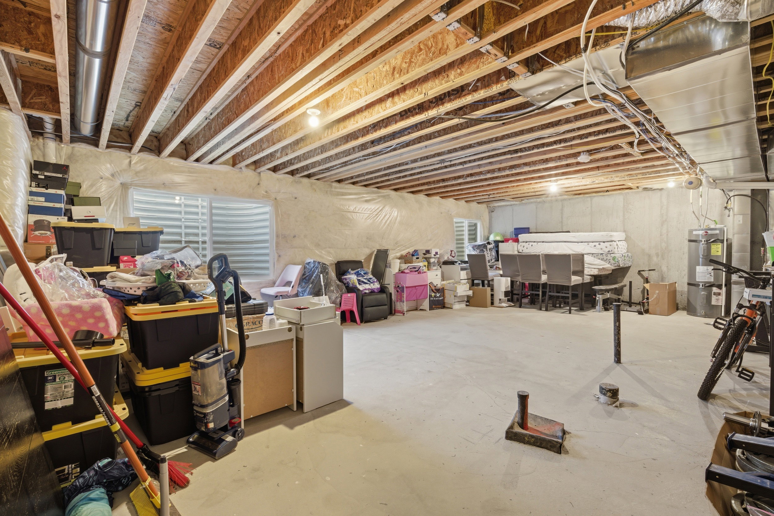 Spacious unfinished basement with exposed wood joists and concrete flooring, featuring an egress window, utilities, and ample open area