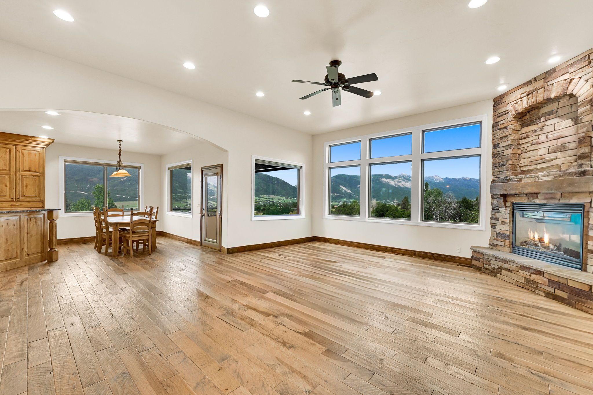 Spacious interior featuring wood-finish flooring, a stone fireplace with a hearth, and a series of large windows showcasing mountain scenery