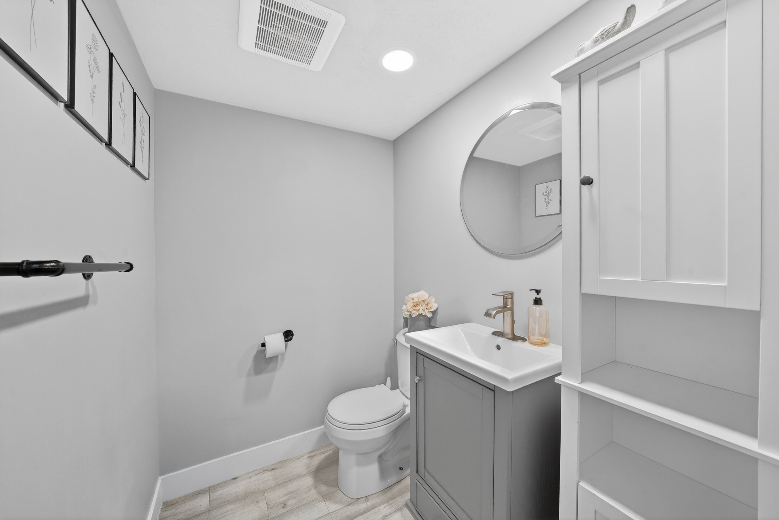Modern powder room featuring a gray vanity with an integrated sink, a circular wall mirror, wood-finish flooring, and a built-in white storage cabinet