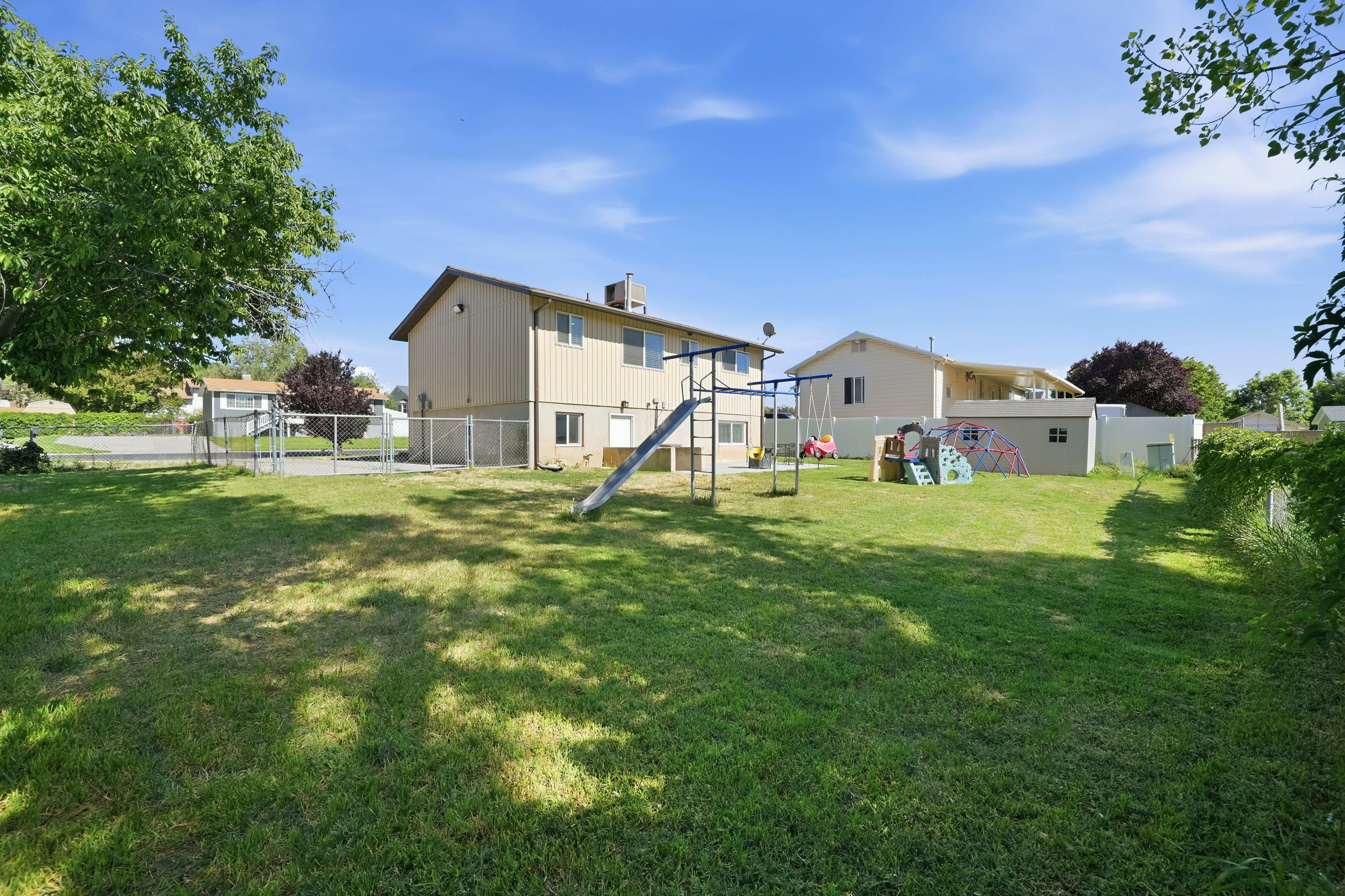 Expansive grassy yard area featuring a chain link fence, mature trees, and a storage shed