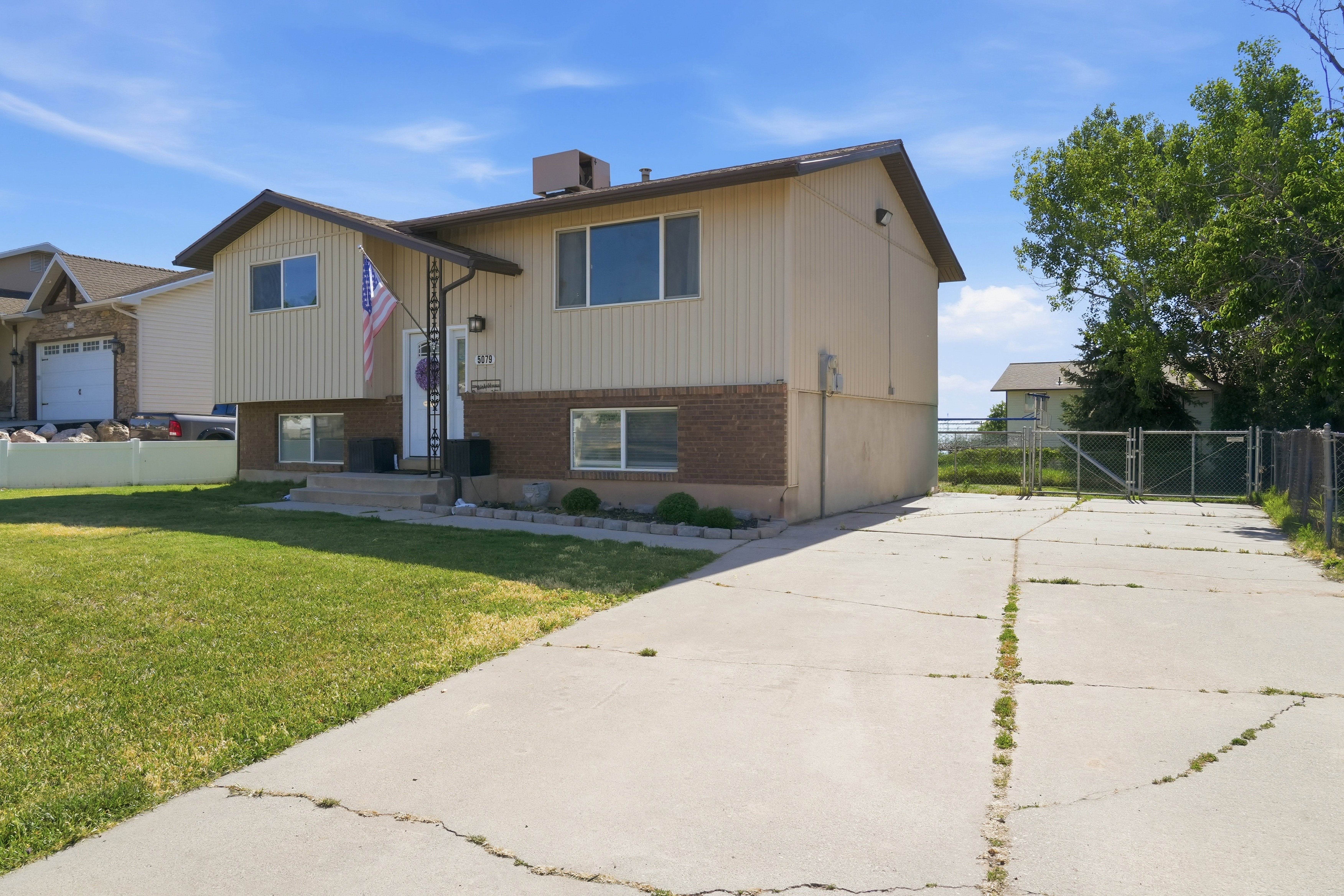 Split-level residence featuring a light-colored siding exterior, brick accent detailing, a covered front entry, a concrete driveway, and an expansive side yard with chain-link fencing