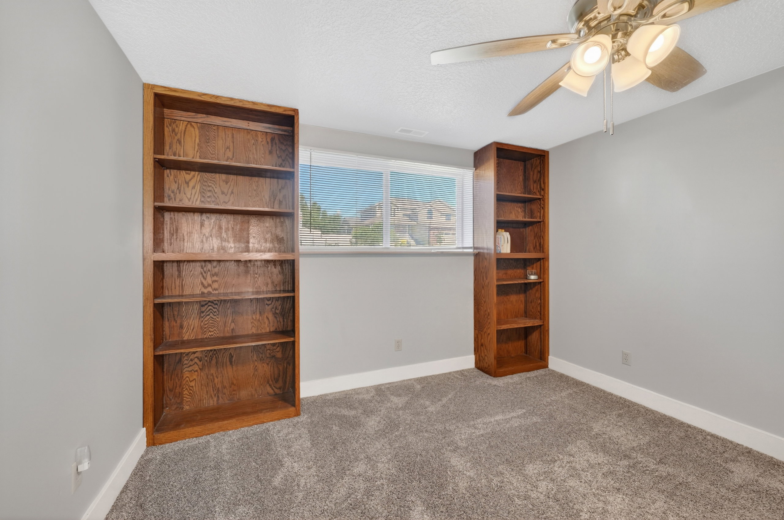 Room featuring two built-in wood-finish bookshelves, a window with horizontal blinds, textured gray carpet, and a ceiling fan with integrated lighting