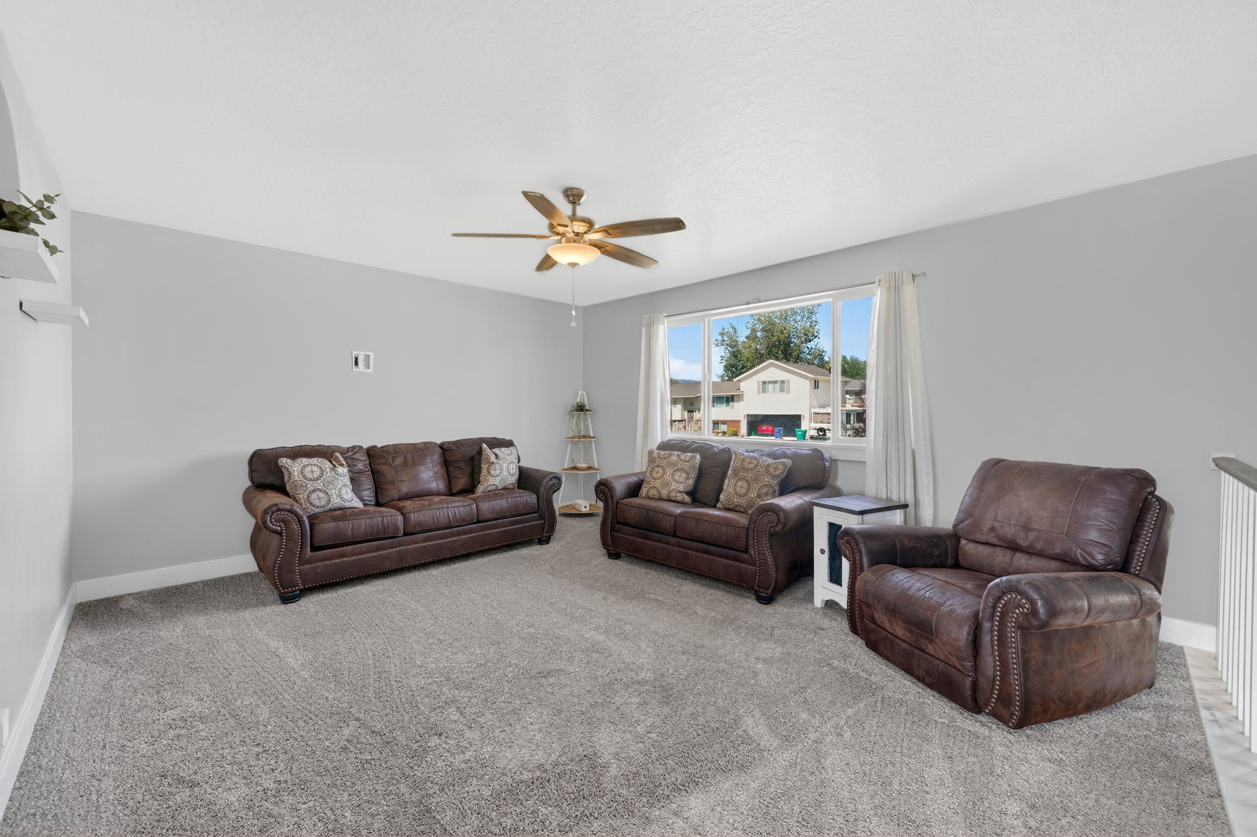 Spacious living area featuring light gray walls, plush gray carpeting, a large window with white trim, and a ceiling fan with integrated lighting