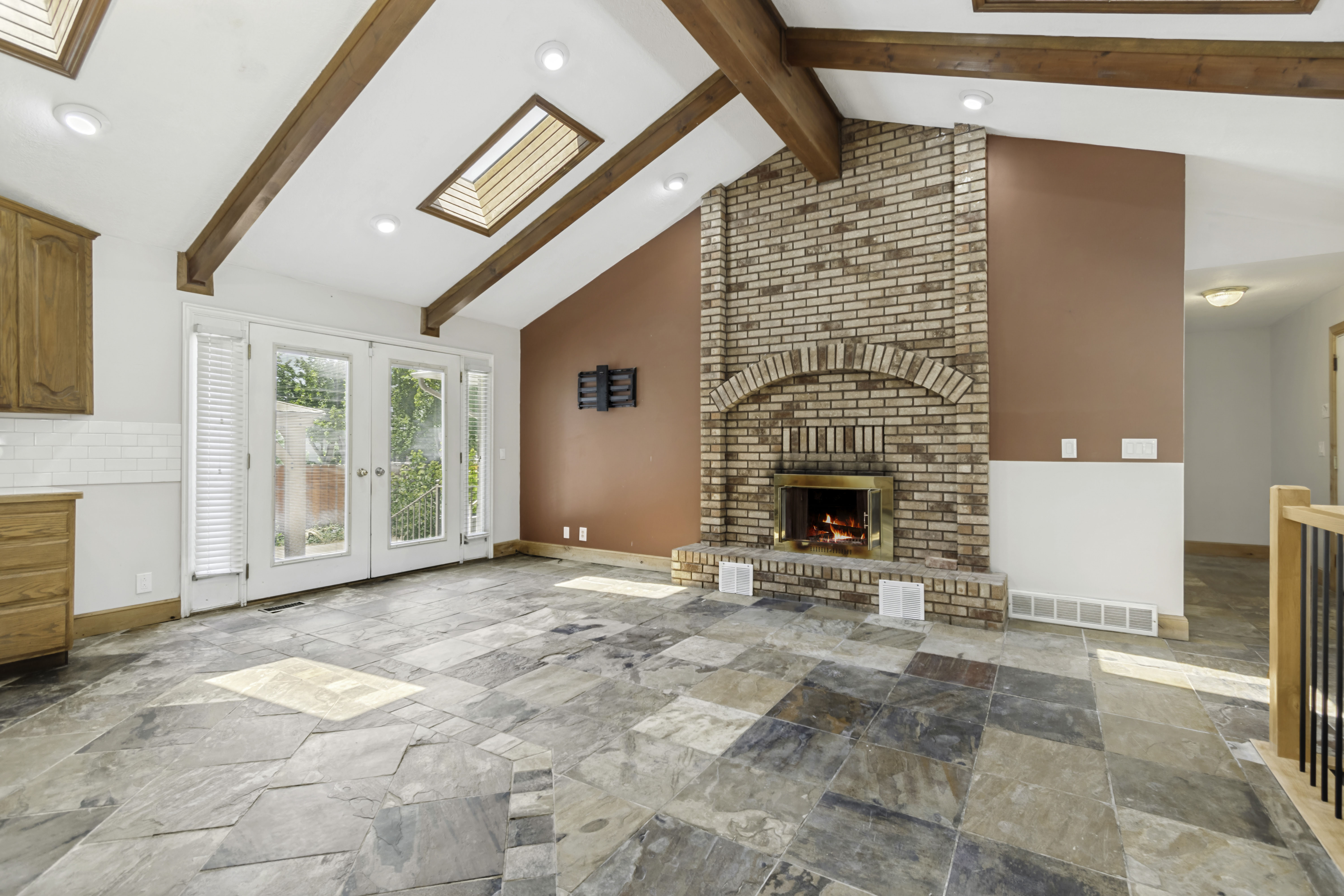Vaulted ceiling with exposed wood beams, brick fireplace with brass firebox, skylights, multi-pane double doors, and natural stone flooring