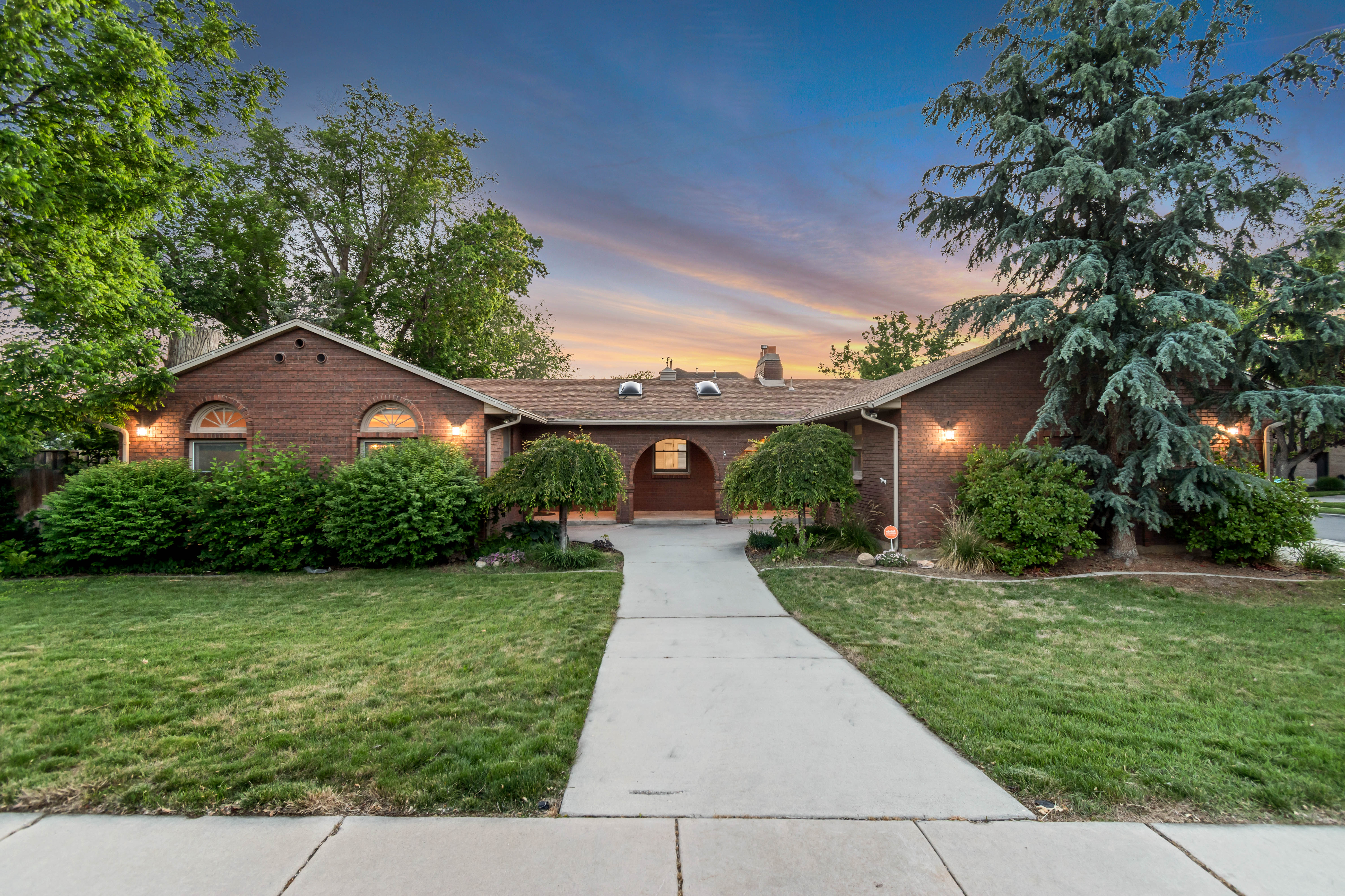 Brick exterior with arched window details, a prominent central archway, mature landscaping, a concrete walkway, and exterior lighting fixtures
