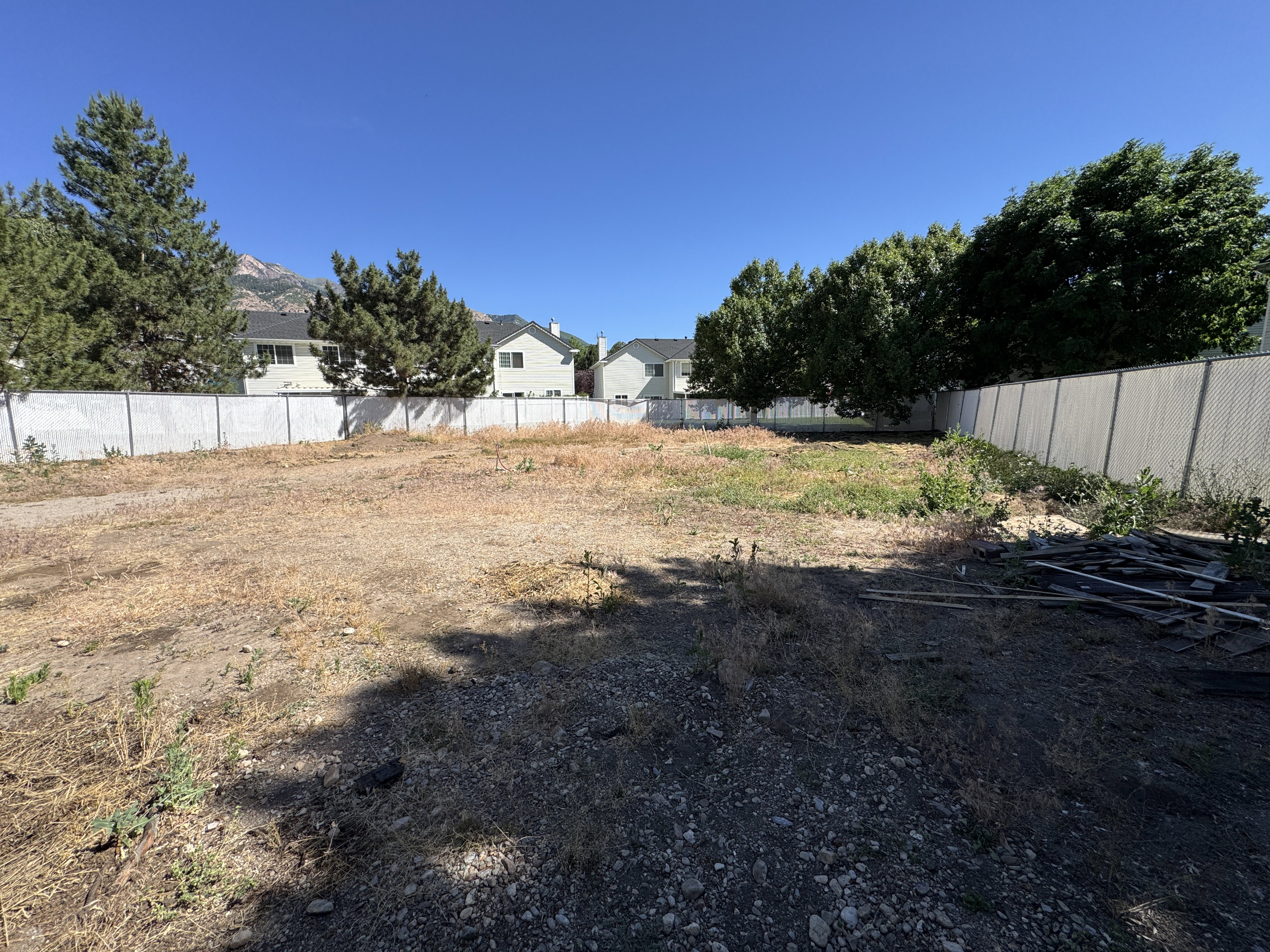 Expansive vacant lot featuring a partial chain-link fence, mature perimeter trees, and mountain scenery