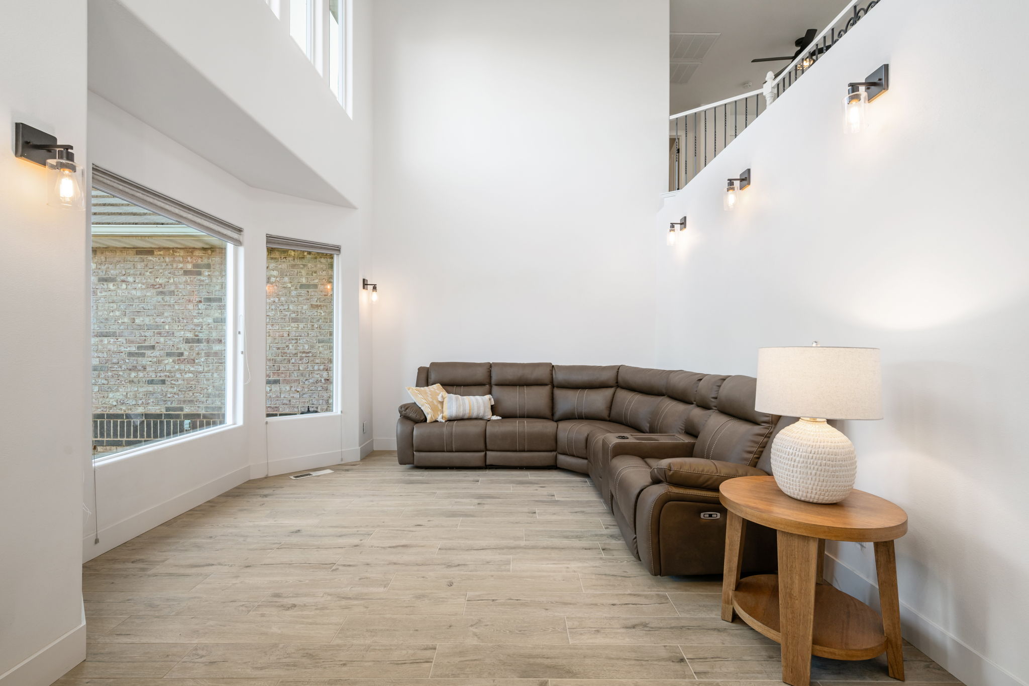Two-story living area with high ceilings, featuring wood-finish flooring, a series of tall windows, and multiple wall-mounted light fixtures
