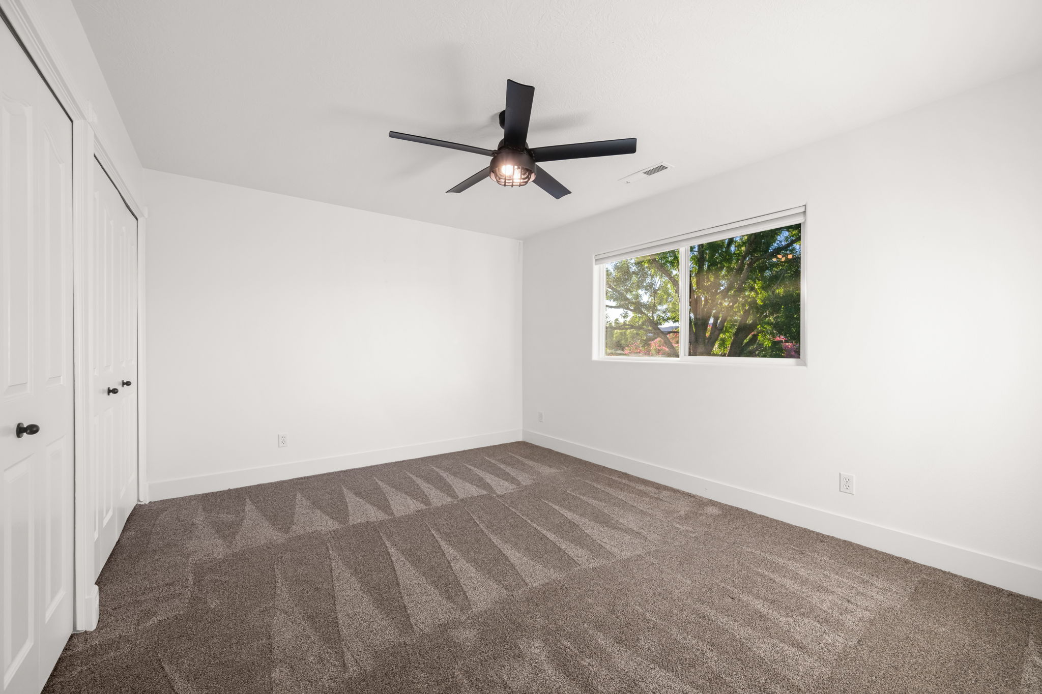 Carpeted room featuring a ceiling fan with light, white walls, and a window overlooking greenery