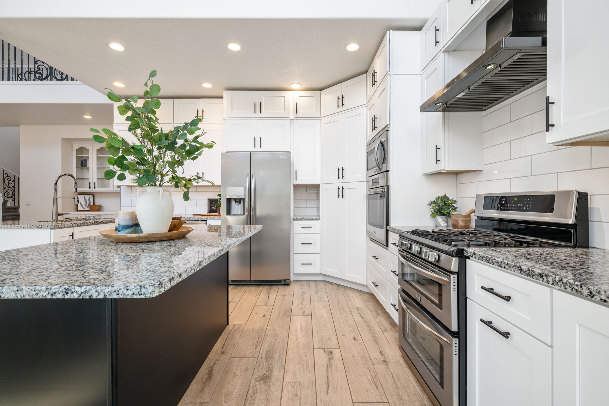 Modern kitchen featuring a large island with a speckled countertop, light wood-finish flooring, white cabinetry with black hardware, stainless steel appliances, and a subway tile backsplash