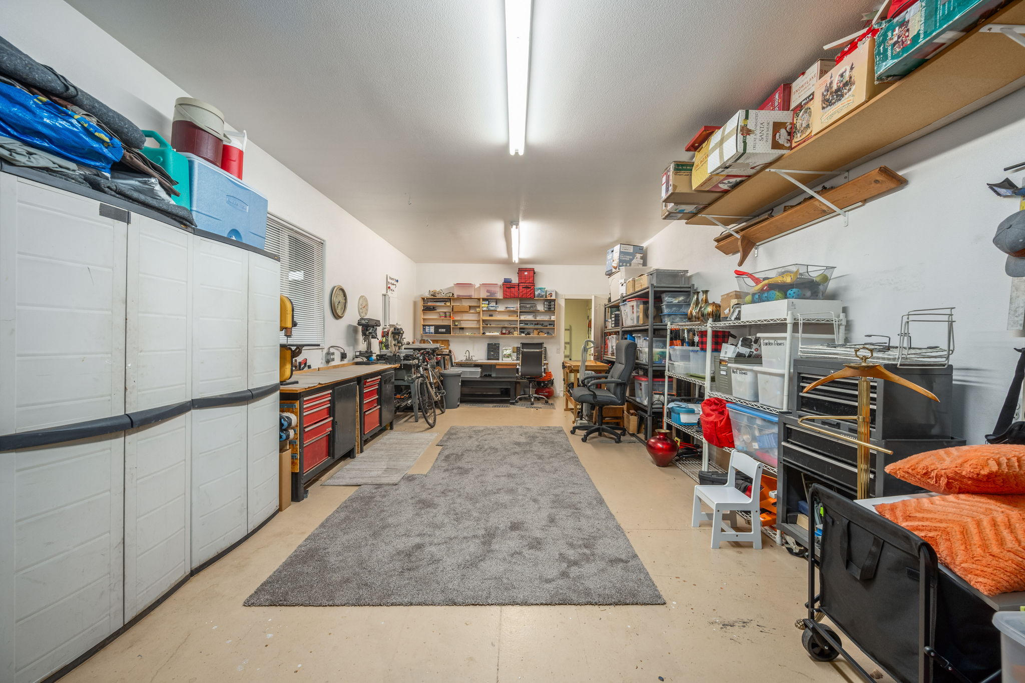 Spacious garage featuring light-colored flooring, abundant built-in shelving, a workbench with integrated cabinetry, and overhead fluorescent lighting