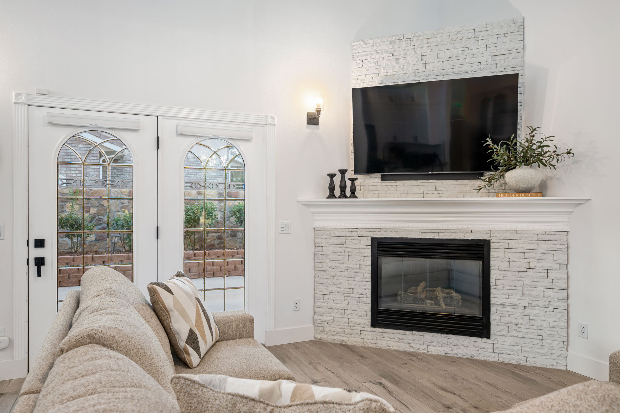 Arched glass panel doors with grid details, a fireplace with a light-toned stacked stone surround, a substantial white mantel, and wood-finish flooring