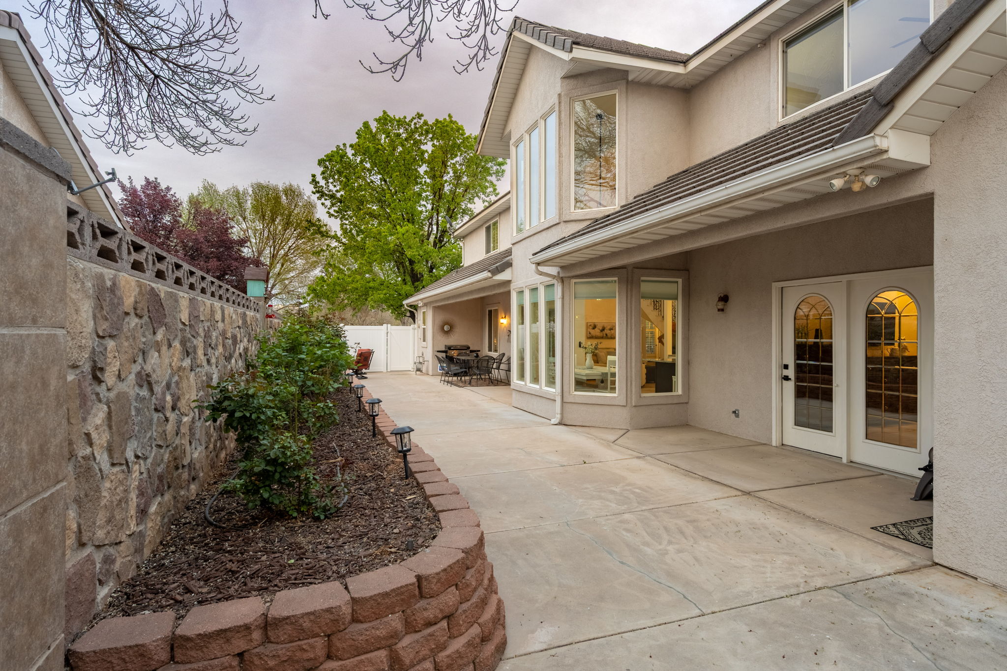 Rear exterior featuring a stone retaining wall, mature landscaping, large windows, and a patio with arched glass doors