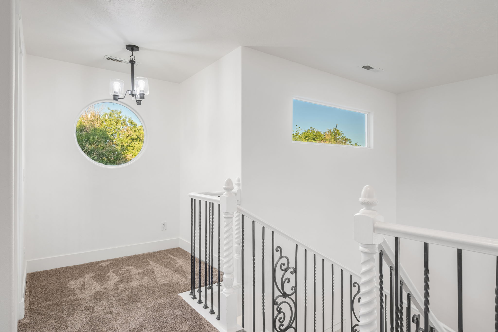 Landing area featuring a distinctive circular window, a rectangular transom window, a black metal chandelier, white balusters with decorative ironwork, and neutral-toned carpeting