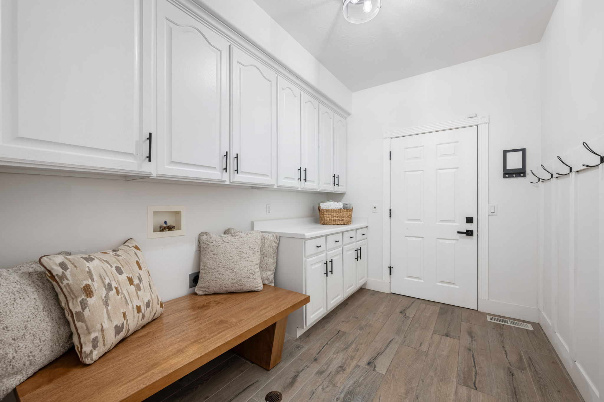 Light-filled utility room featuring extensive white cabinetry with matte black hardware, a wood-finish bench, a built-in countertop, and wood-finish flooring