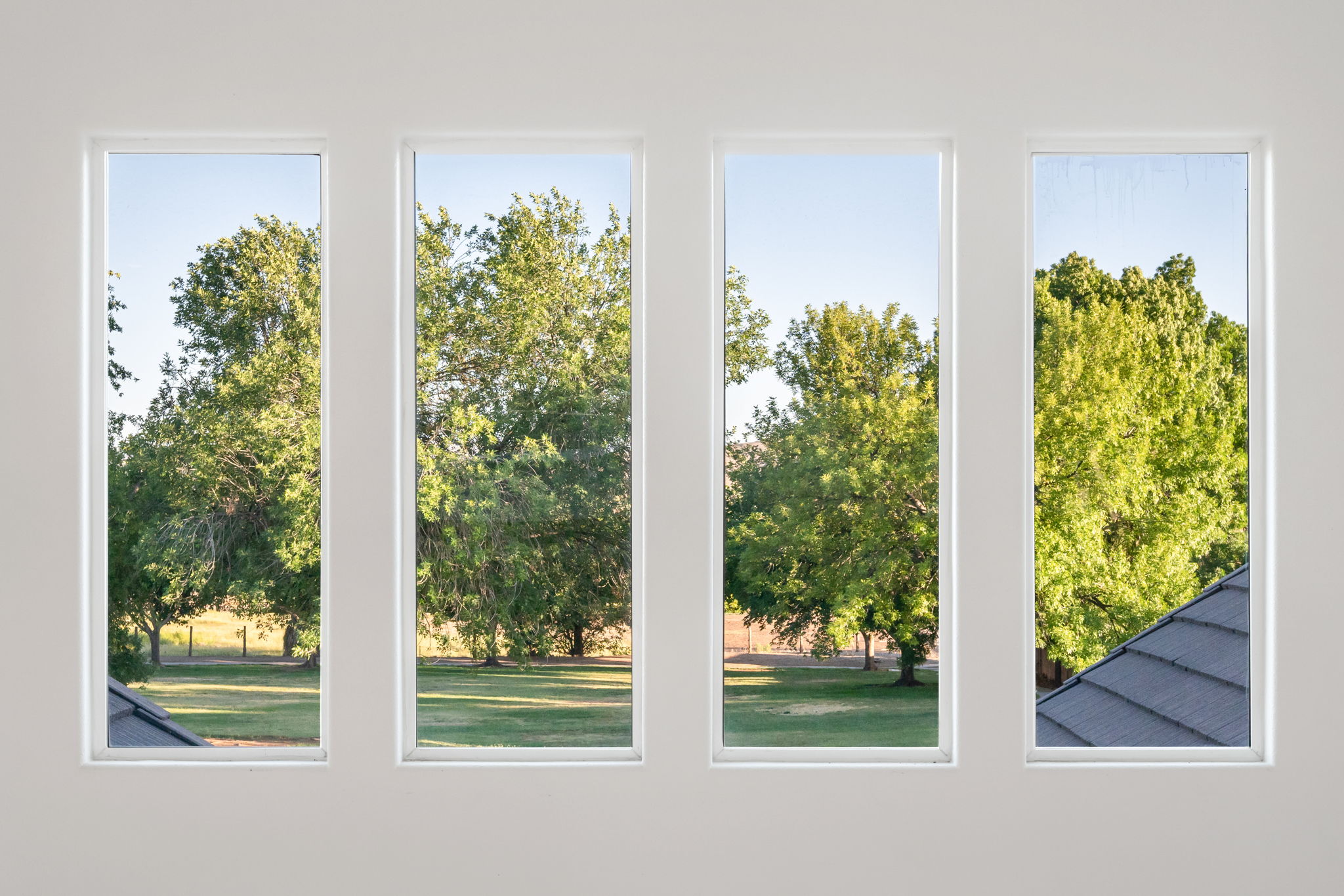 Four vertical windows with white frames overlooking a green landscape with mature trees and a partial dark-tiled roof