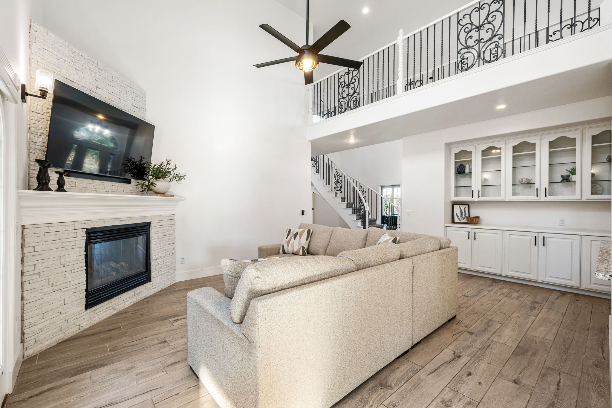 Living area featuring a soaring vaulted ceiling, stone-clad fireplace with mantel, wood-finish flooring, custom built-in cabinetry with glass-front uppers, and an open balcony with decorative wrought iron railing