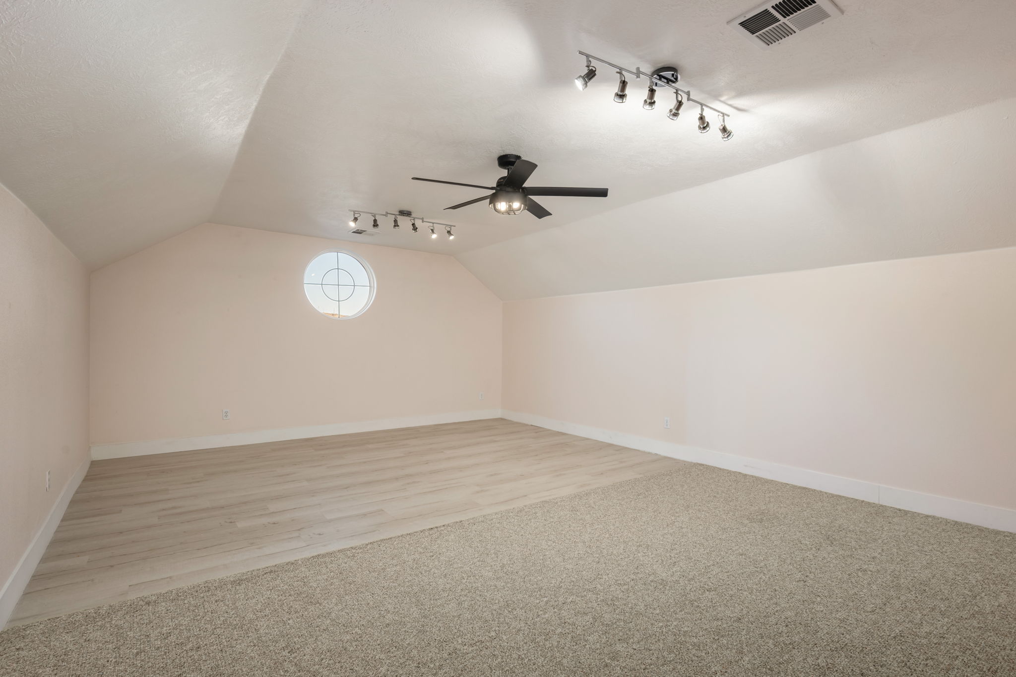 Spacious room featuring a round architectural window, sloped ceilings, track lighting fixtures, and a ceiling fan