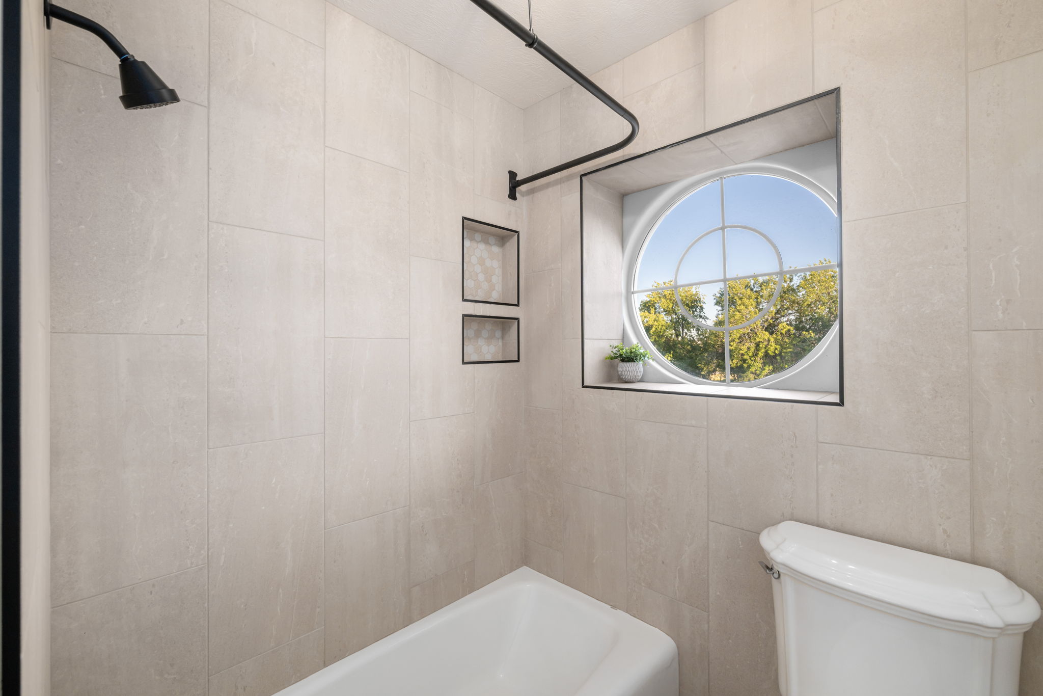 Tub and shower combination featuring floor-to-ceiling light-toned tile, a circular window with black trim, and a black shower fixture