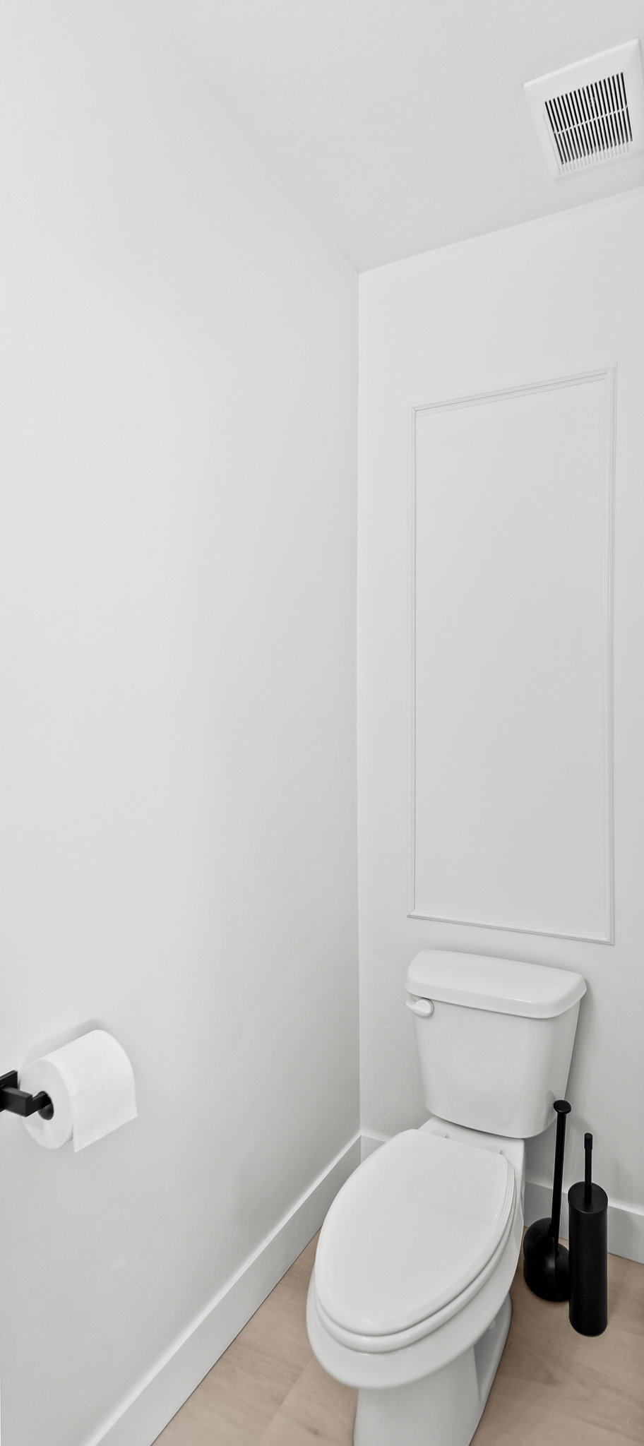White toilet with a built-in panel accent wall, light wood-finish flooring, and a black wall-mounted toilet paper holder