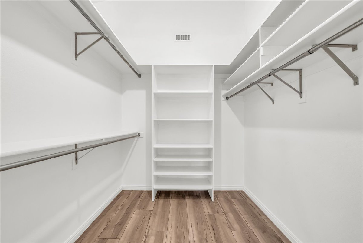 Walk-in closet featuring wood-finish flooring, white walls, built-in shelving, and multiple clothing rods with supporting brackets